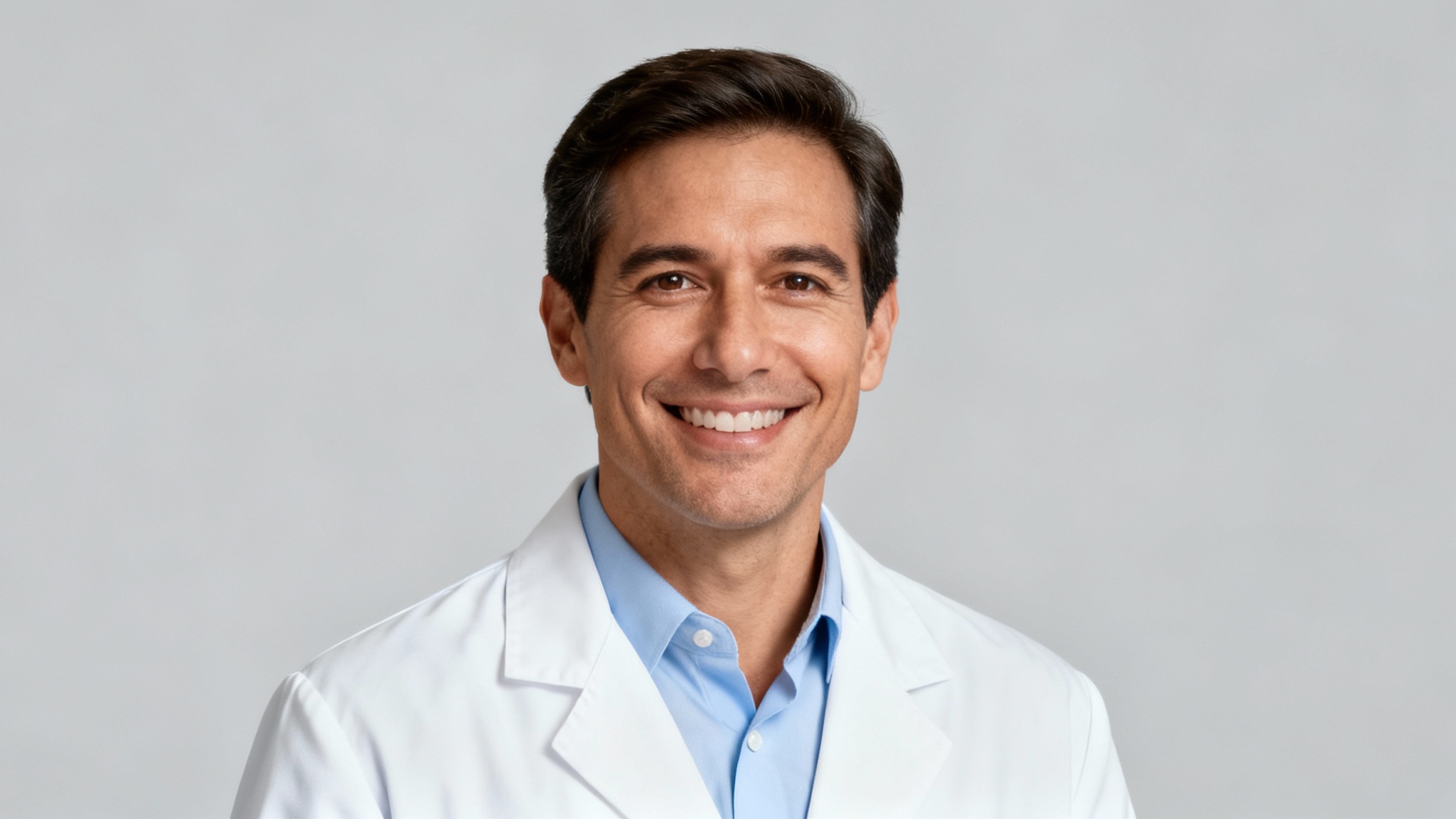 A professional headshot of a smiling male doctor wearing a white coat, looking directly at the camera against a plain light gray background.