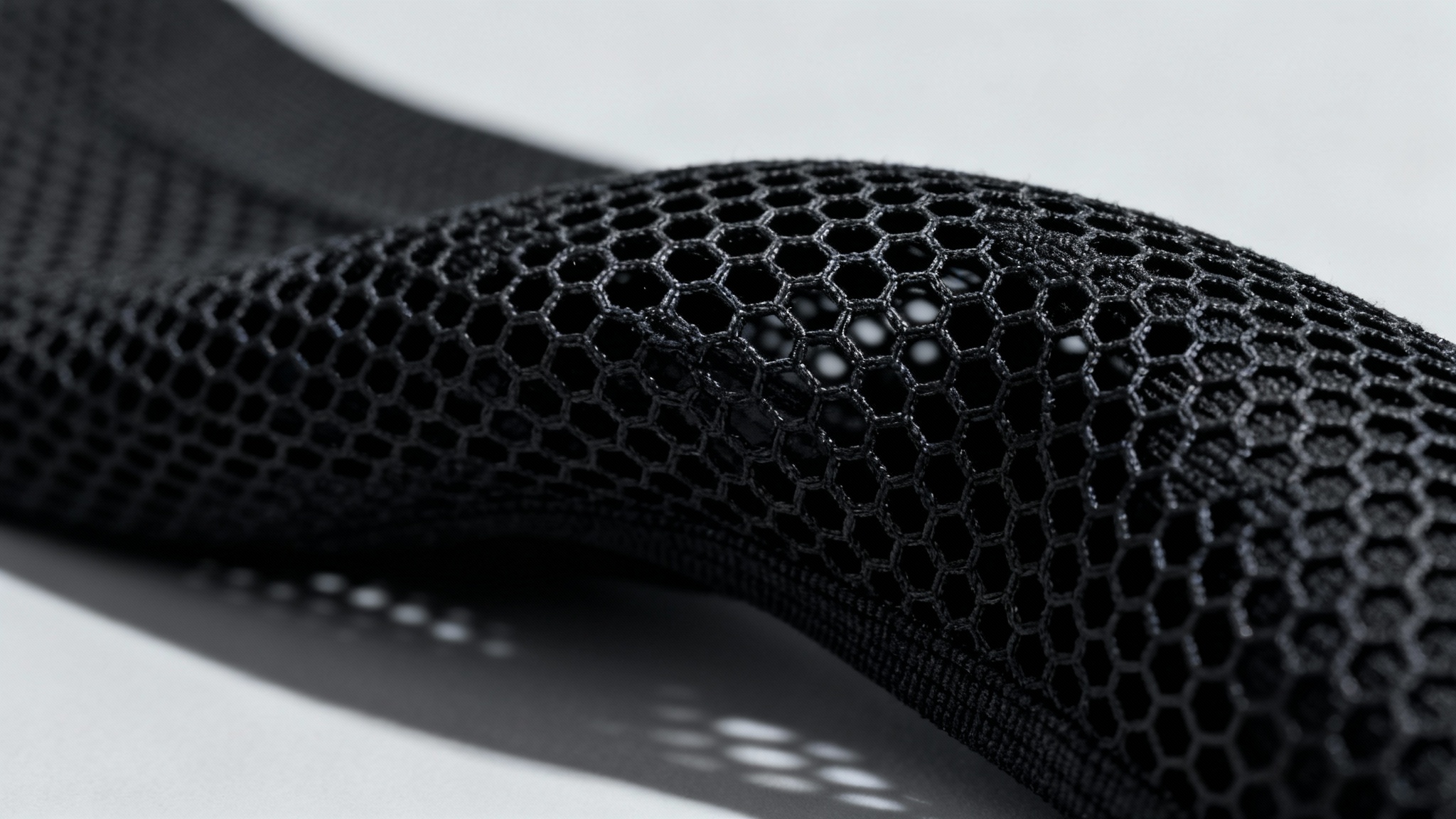A detailed macro photograph of a black technical mesh fabric, showcasing the intricate hexagonal weave and texture of the material under dramatic studio lighting.