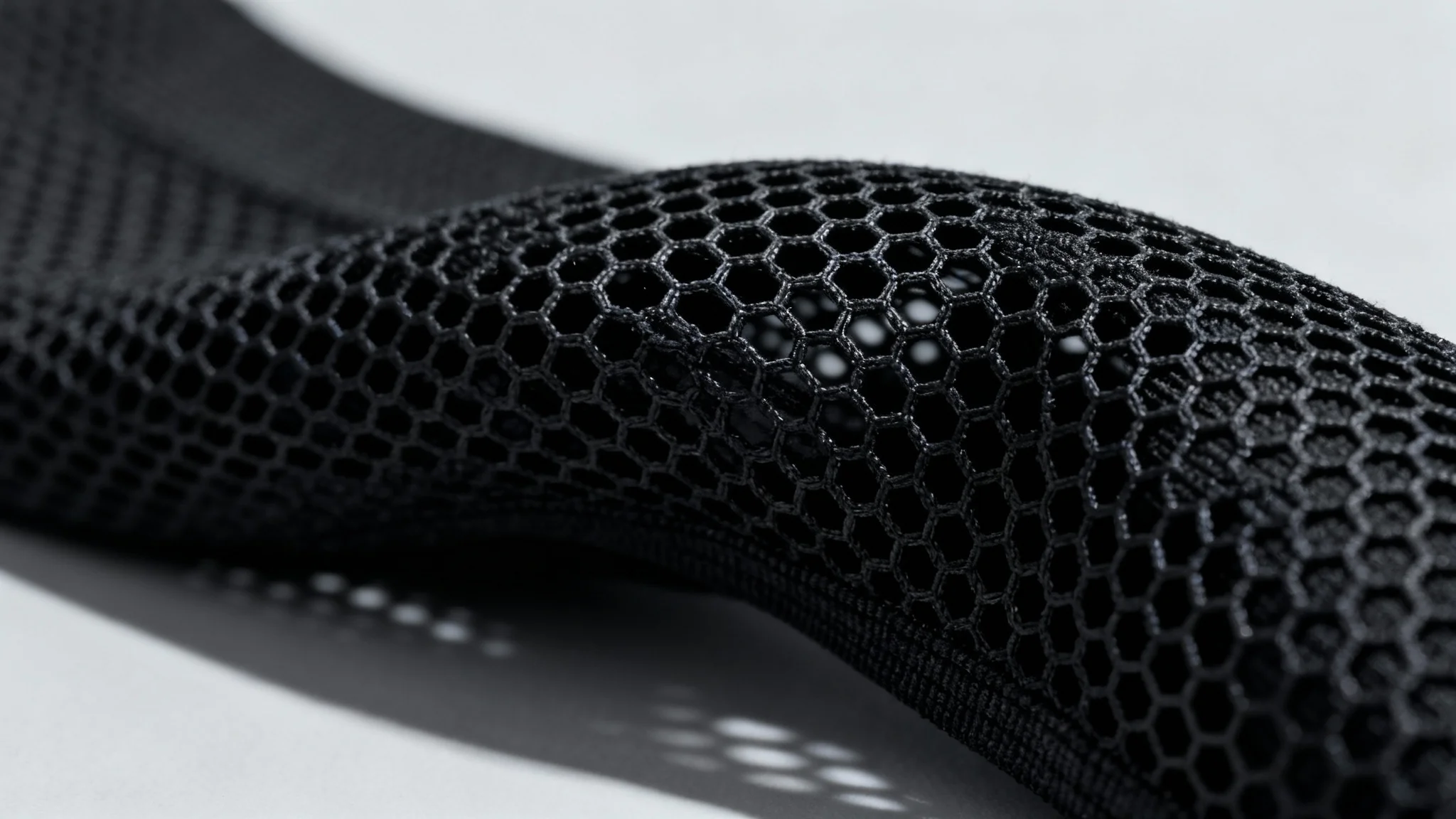 A detailed macro photograph of a black technical mesh fabric, showcasing the intricate hexagonal weave and texture of the material under dramatic studio lighting.