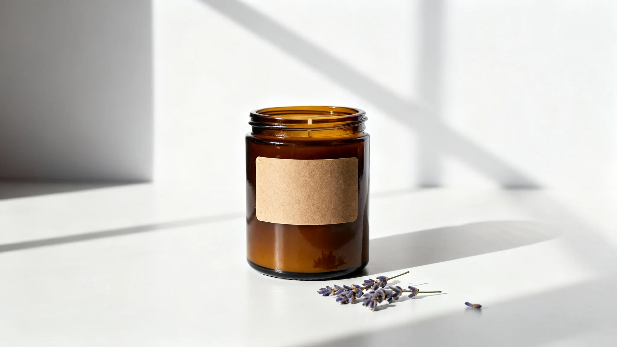 A professional product photo of a handmade scented candle in an amber glass jar, placed on a white surface with dried lavender sprigs, against a clean white background.