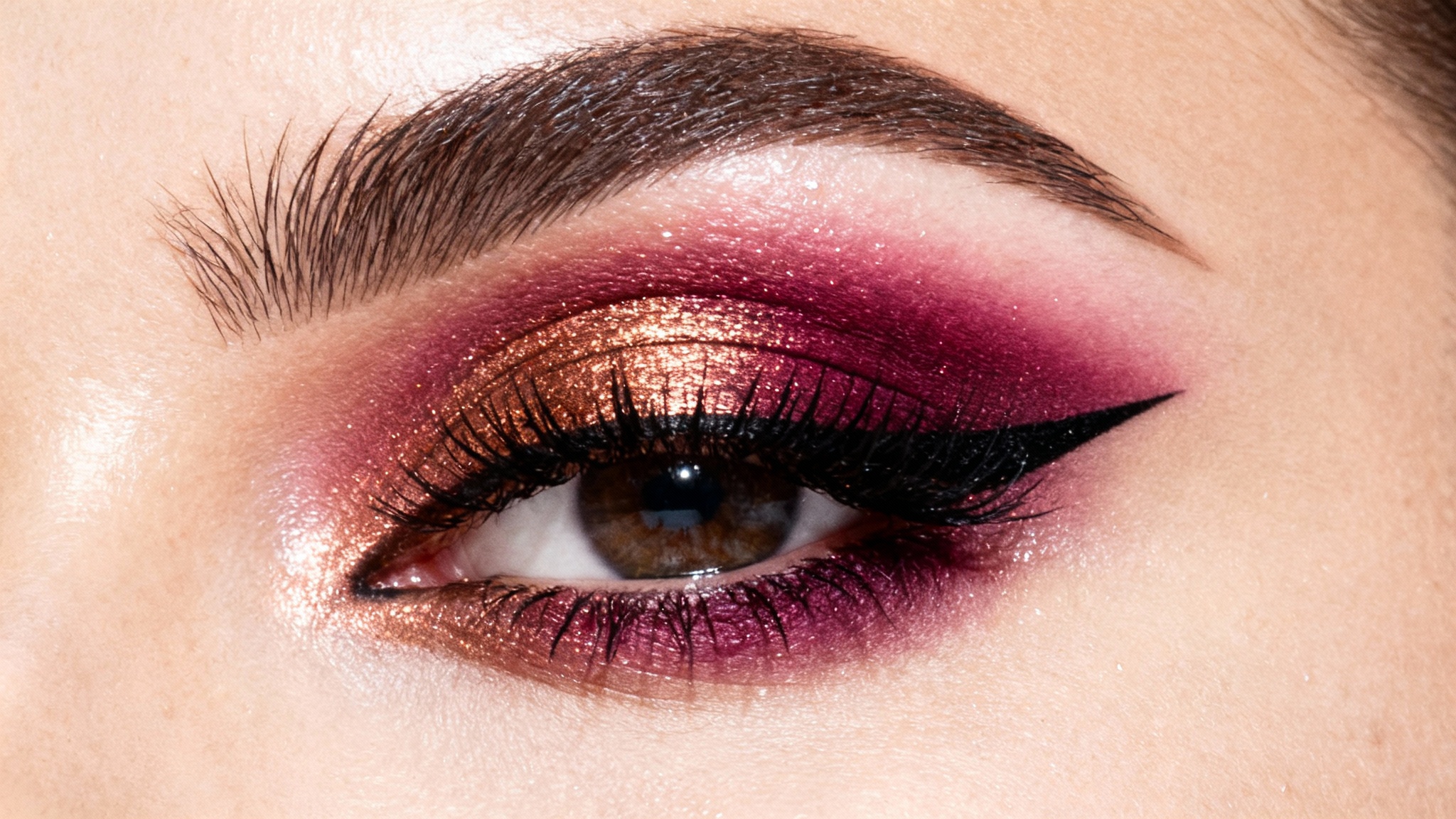 An extreme close-up of a woman's eye showcasing perfectly applied copper and magenta eyeshadow, bold winged eyeliner, and long lashes against a plain white background.