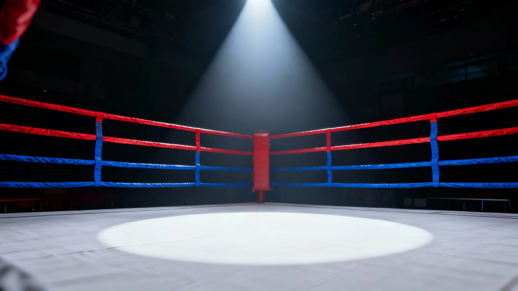 A dramatic, cinematic photo of an empty boxing ring illuminated by a single spotlight from above, with taut red and blue ropes, set against a dark, empty background.