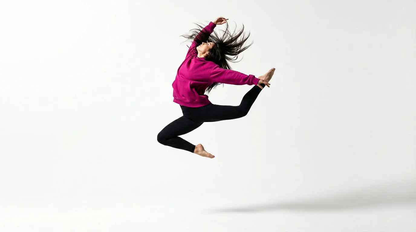 A still frame from a vertical video showing a female dancer in a bright pink hoodie captured in mid-air during a dynamic leap against a clean white studio background.