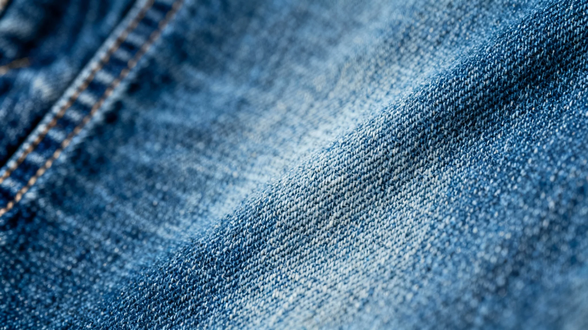 A detailed, high-quality close-up image of a blue jeans denim texture, suitable for use as a background. The diagonal weave and individual threads are clearly visible under soft lighting.
