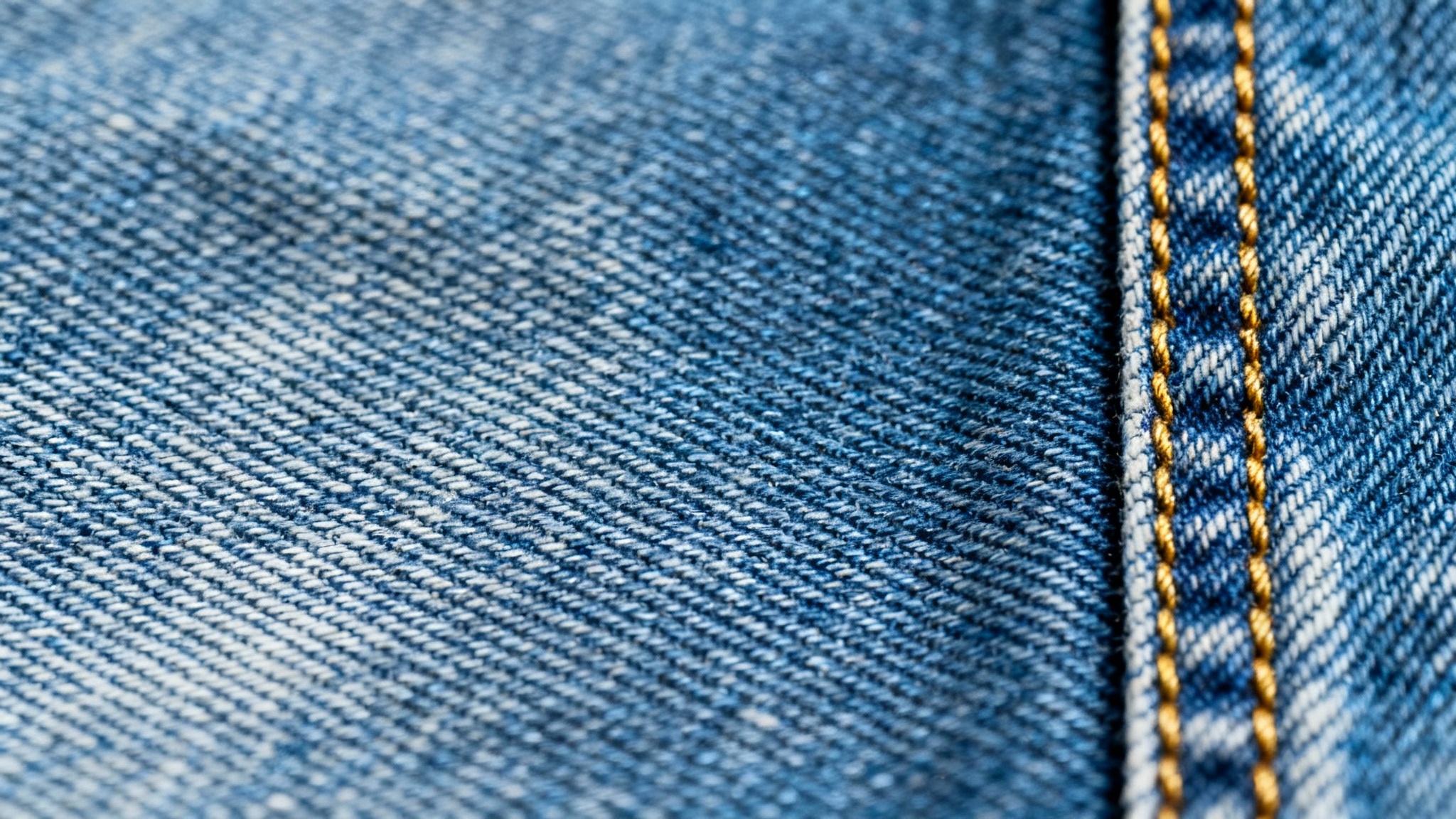 A detailed macro shot of blue jeans fabric, showing the classic denim texture and a vertical seam with gold-colored stitching on a white background.