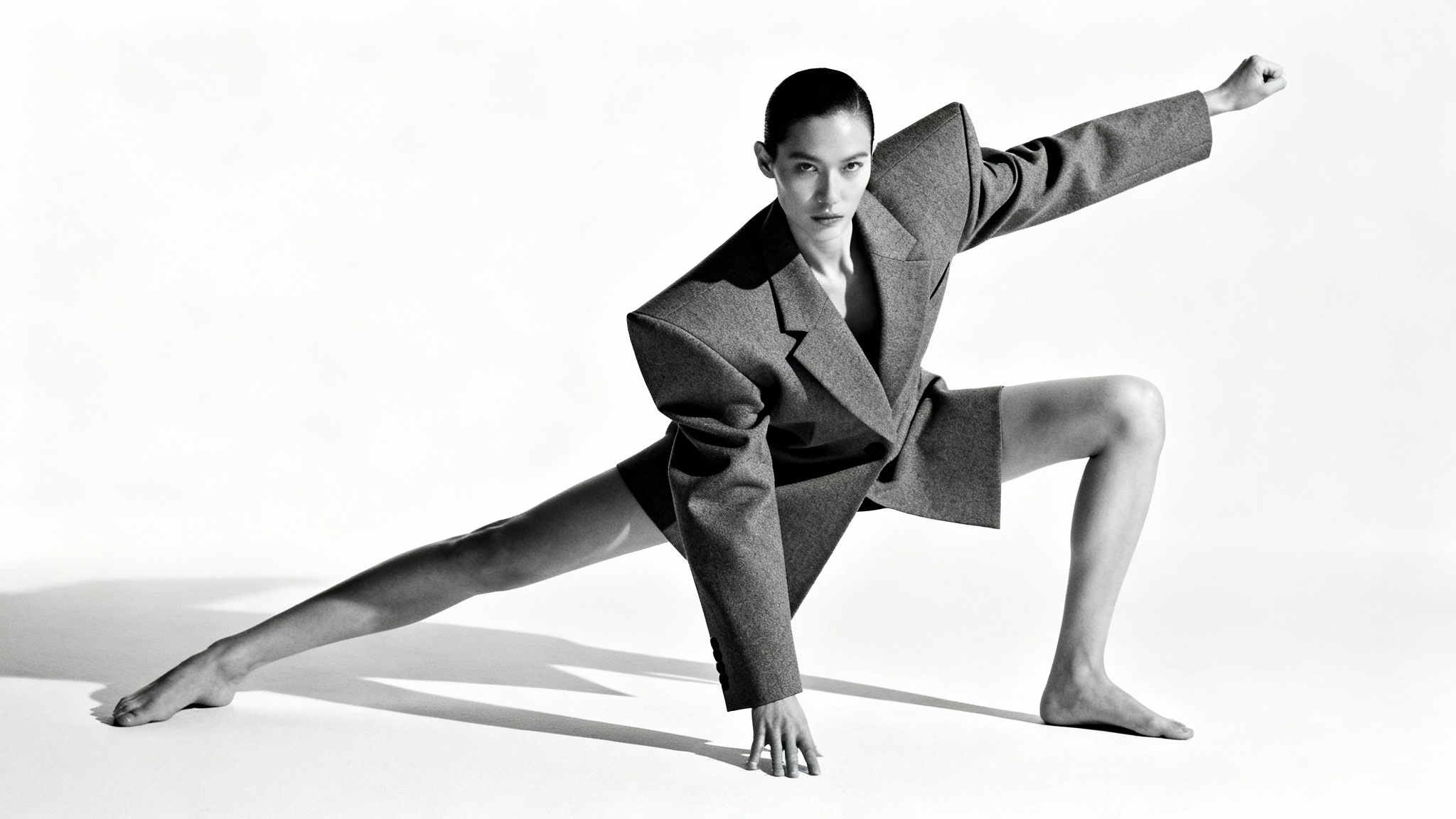 A full-body shot of a high fashion model in a dramatic, angular pose, wearing a structural grey outfit against a stark white background.