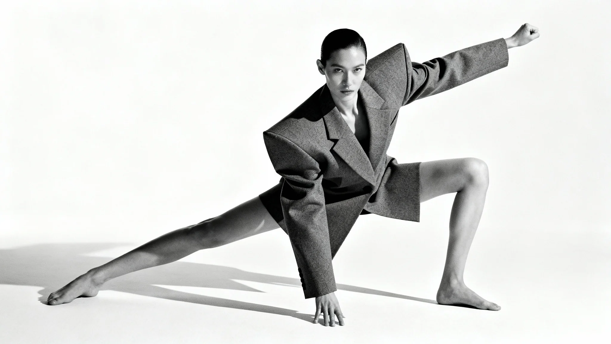 A full-body shot of a high fashion model in a dramatic, angular pose, wearing a structural grey outfit against a stark white background.