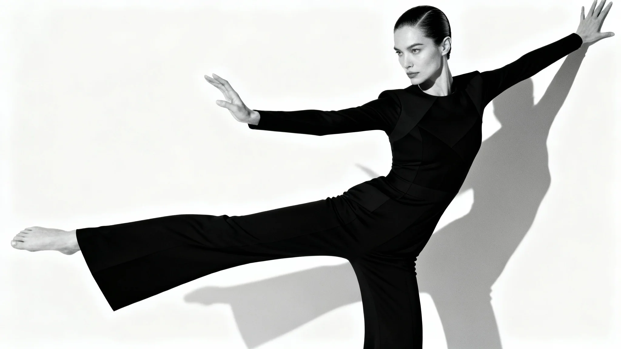 A fashion model in a sleek black jumpsuit strikes a dramatic, angular pose against a stark white background in a photo studio.