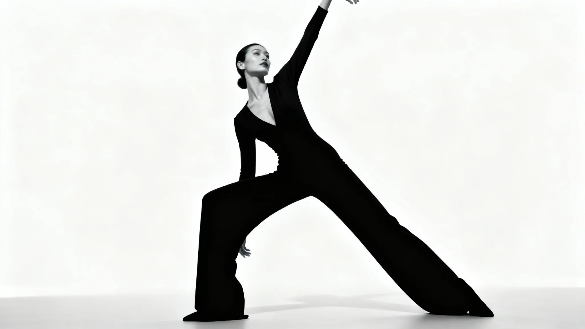 A full-body photograph of a female model in a dynamic pose, wearing a black jumpsuit against a stark white studio background, captured with dramatic lighting.