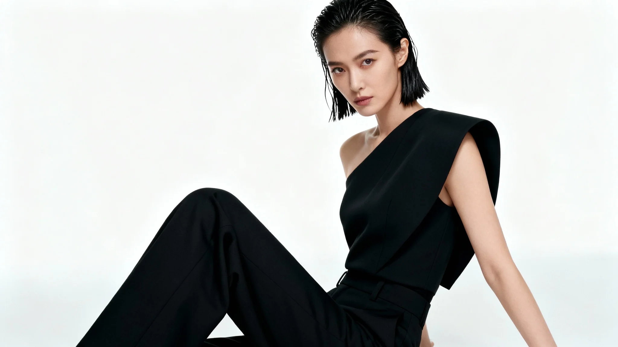 A full-body photograph of a high fashion model with dark hair posing confidently in a stylish, minimalist black outfit against a plain white studio background.