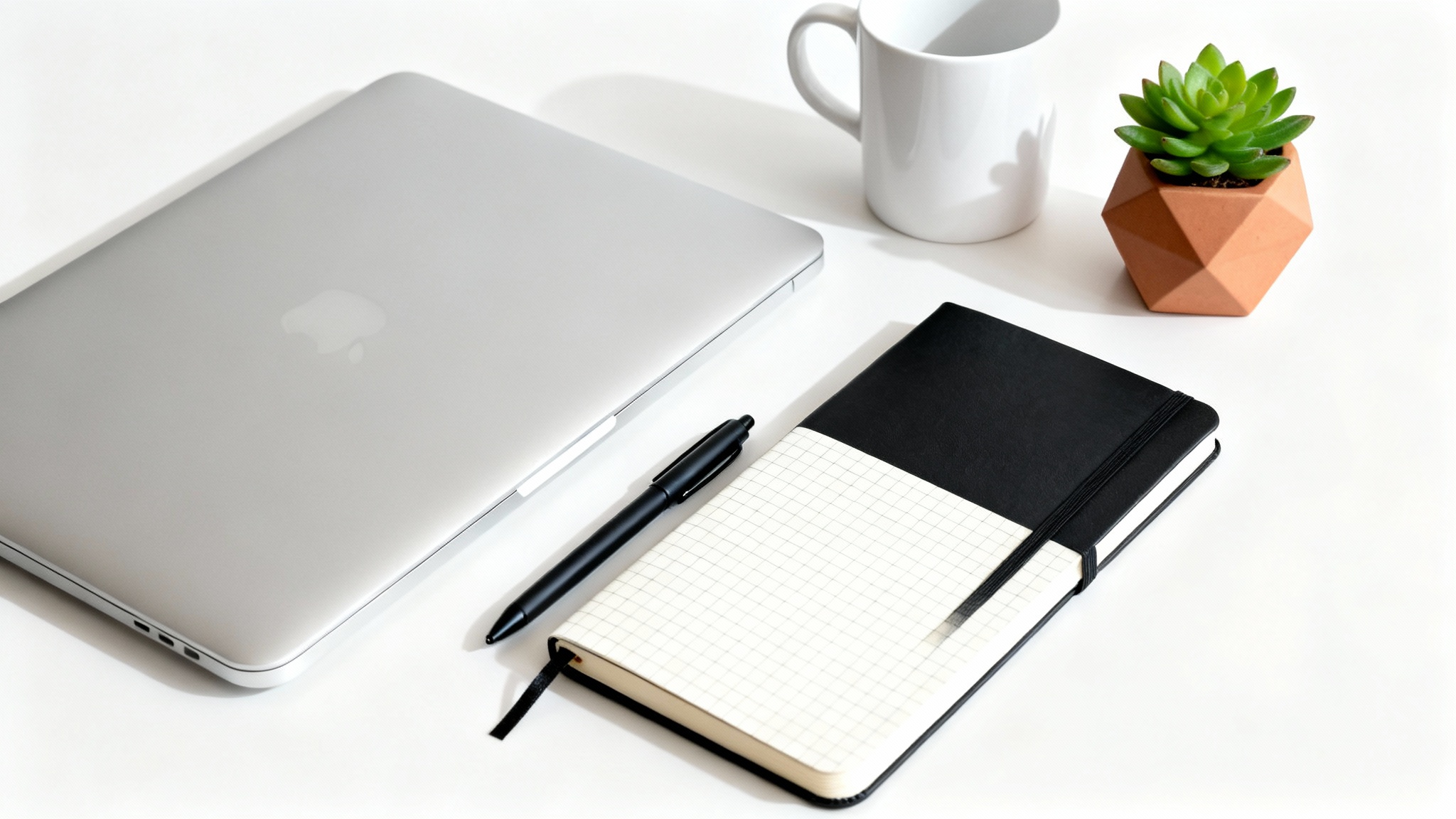 A top-down view of an organized desk with a closed laptop, a notebook, a pen, a coffee mug, and a small plant, on a white background.