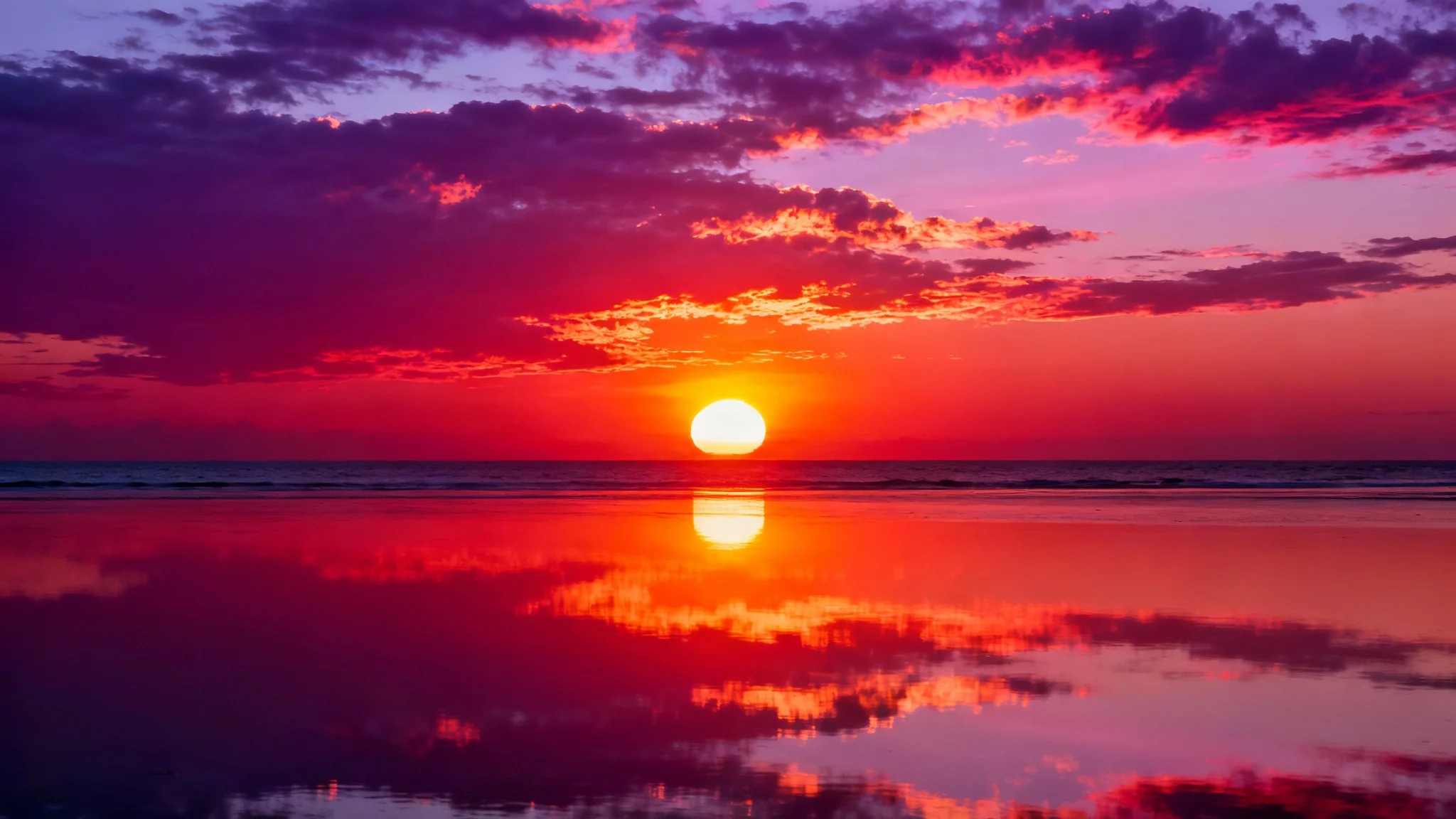 A photorealistic image of a vibrant sunset over a calm ocean, with the sky filled with dramatic shades of orange, pink, and purple, all reflected in the water below.